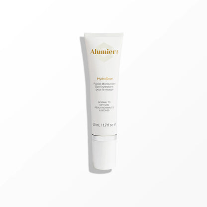 A white cylindrical bottle with a silver cap labeled "Alumier HydraDew Facial Moisturizer for normal to dry skin, 50 mL / 1.7 fl oz," stands upright on a white surface against a pale background.