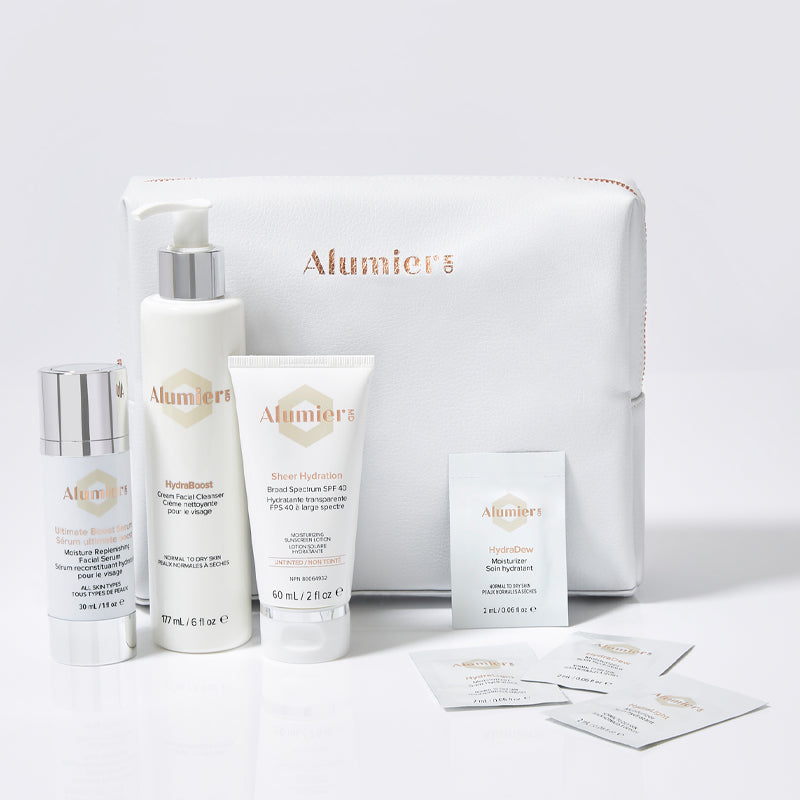 A set of Alumier skincare products, including pump bottle, tube, and small packets, arranged in front of a white cosmetic bag with the Alumier logo, on a white background.