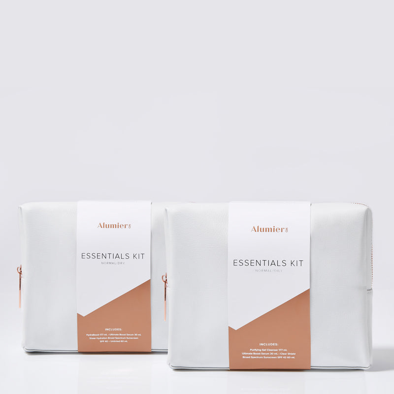 Two white pouches with rose gold zippers sit against a plain background. Each pouch has a label reading "AlumierMD ESSENTIALS KIT" followed by "INCLUDES" and a list of contents at the bottom.