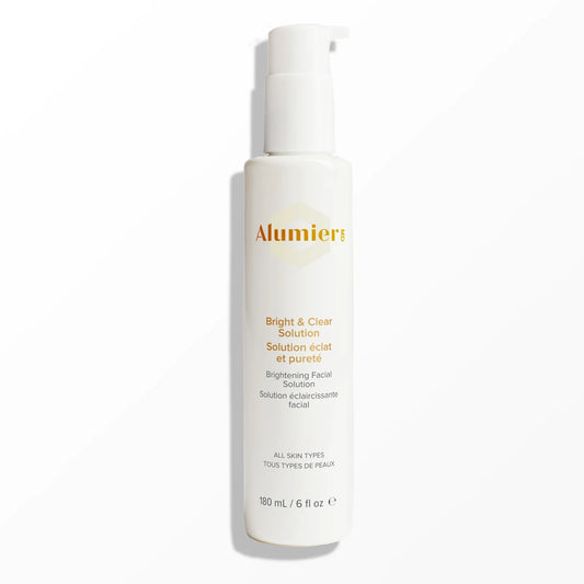 A white bottle labeled "Alumier" contains Bright & Clear Solution, a brightening facial product, placed on a plain white surface. Text reads: "All skin types" and "180 ml / 6 fl oz."