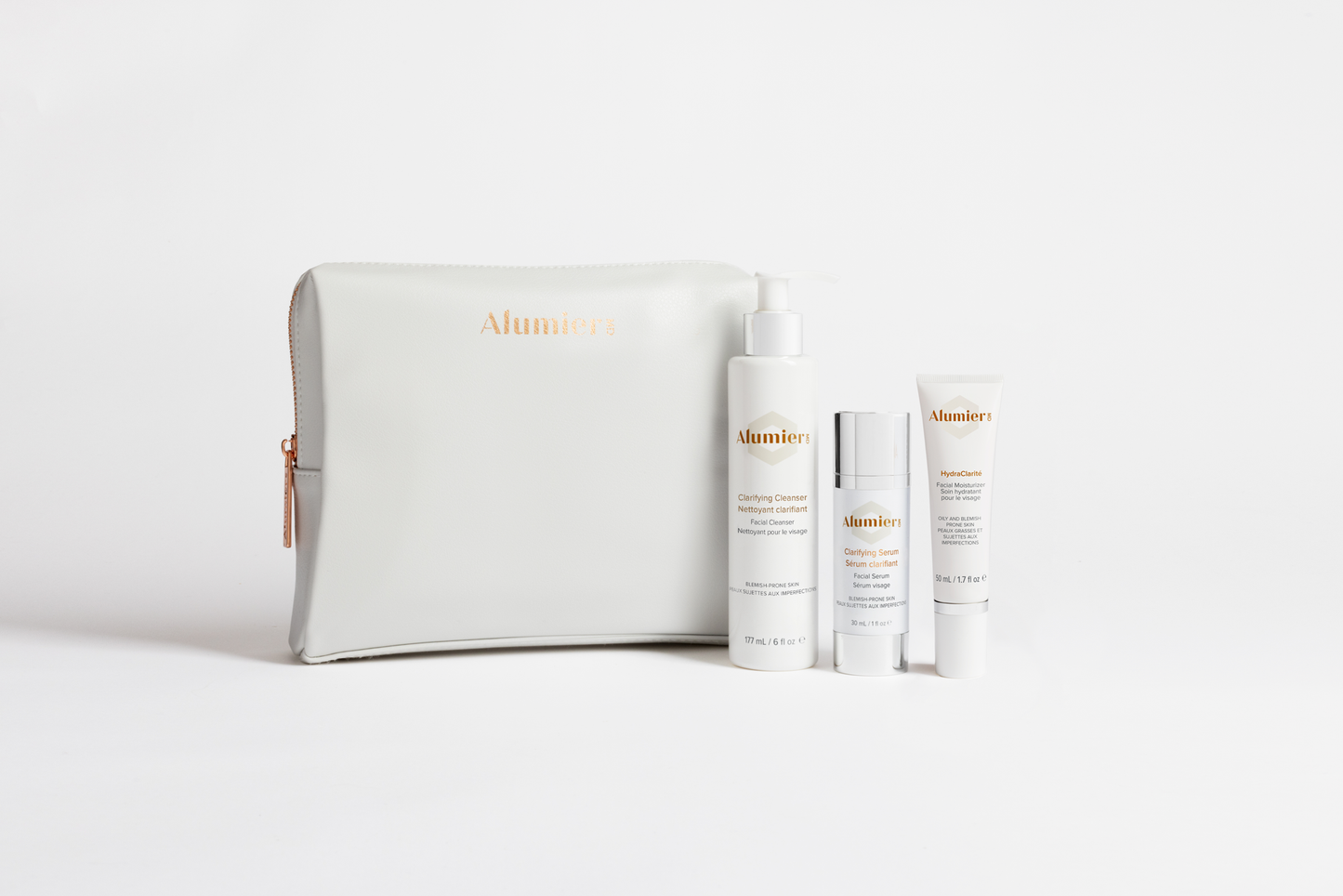 A set of AlumierMD skincare products, including a cleanser, serum, and sunscreen, stand upright next to a zippered cosmetic bag against a white background.