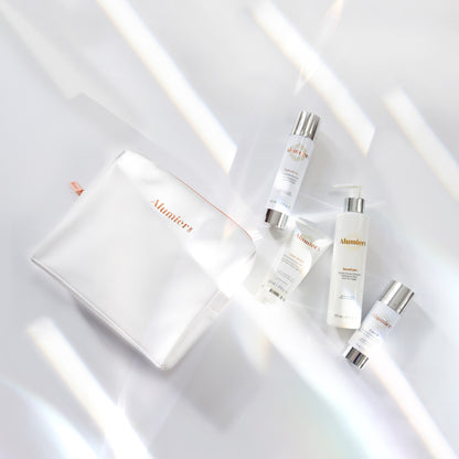 Skincare products with silver caps lay beside a white cosmetic bag, marked "Alumier," arranged on a white background with soft light reflections. Product descriptions are partially visible on the bottles.