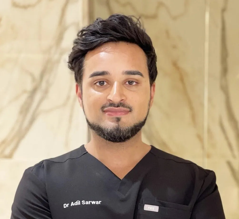 A man in a black medical uniform stands facing forward, smiling slightly, in a room with a marbled wall background. Text on the uniform reads, "Dr Adil Sarwar," and "FIGS."