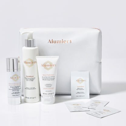 A set of Alumier skincare products, including pump bottle, tube, and small packets, arranged in front of a white cosmetic bag with the Alumier logo, on a white background.