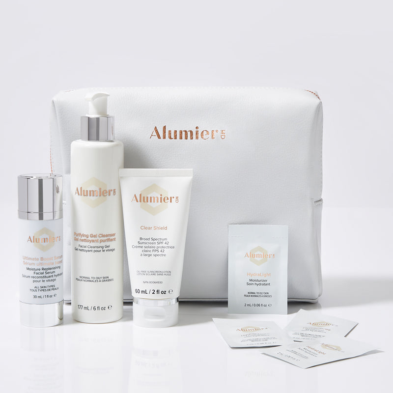 Skincare products from AlumierMD, including bottles, tubes, packets, and a white branded zippered pouch. The text reads "AlumierMD," "Ultimate Boost Serum," "Purifying Gel Cleanser," "Clear Shield," and "Hydralight Moisture Mask."