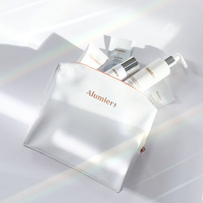 A white pouch labeled "Alumier" holds skincare products, including a pump bottle and tubes. The scene is brightly lit with light streaks, creating a clean, serene atmosphere.