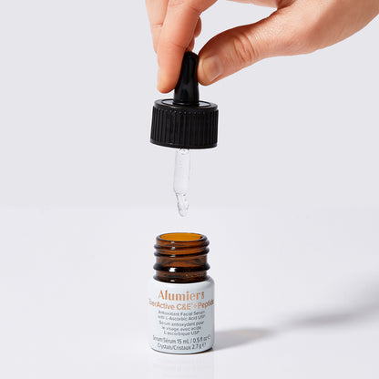 A hand uses a dropper to extract liquid from a brown bottle labeled "AlumierMD EverActive C&E™ + Peptide Antioxidant Facial Serum with L-Ascorbic Acid (USP)." The bottle is placed on a white surface.