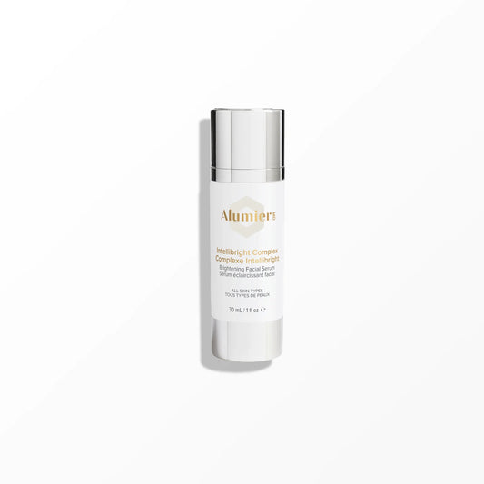 A white cylindrical bottle with a silver cap labelled "Alumier MD Intellibright Complex Brightening Facial Serum, All Skin Types, 30mL / 1.1 fl oz" stands on a smooth, light-grey surface against a plain background.