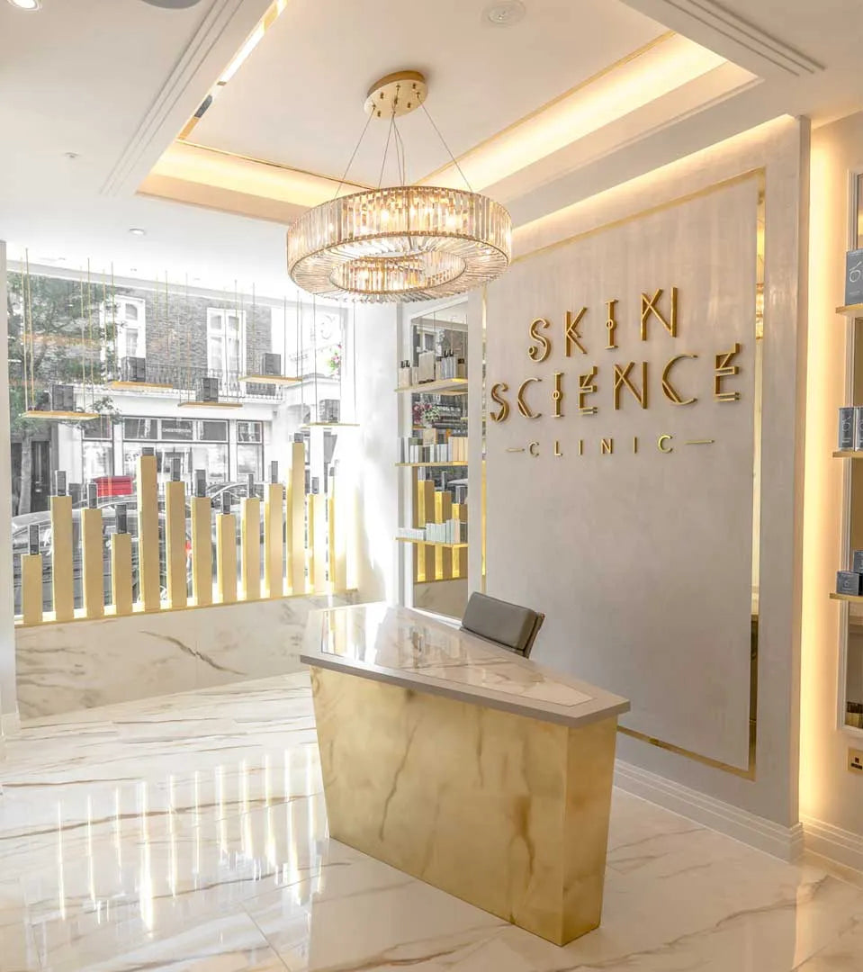 A sleek, modern reception desk stands beneath a crystal chandelier in a spacious, marble-floored clinic. Large golden letters on a wall spell "SKIN SCIENCE CLINIC"; a street view is visible outside.