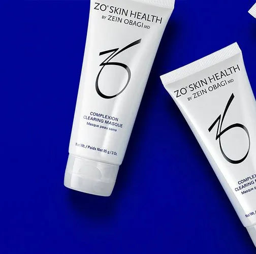 Two white tubes labeled "ZO® SKIN HEALTH BY ZEIN OBAGI MD COMPLEXION CLEARING MASQUE," lying on a blue surface. The text indicates their purpose and branding.