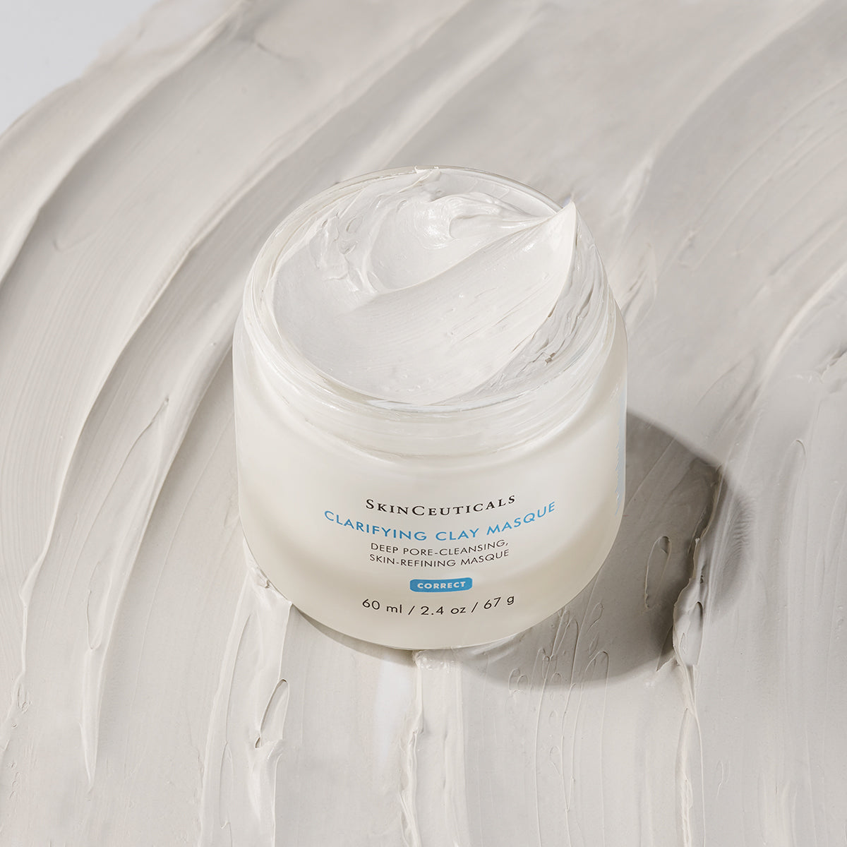 A jar labeled "SkinCeuticals Clarifying Clay Masque" sits open, with creamy white masque inside. The jar is placed on a background of smeared, similar-colored clay. Text: "60 ml / 2.4 oz / 67 g".