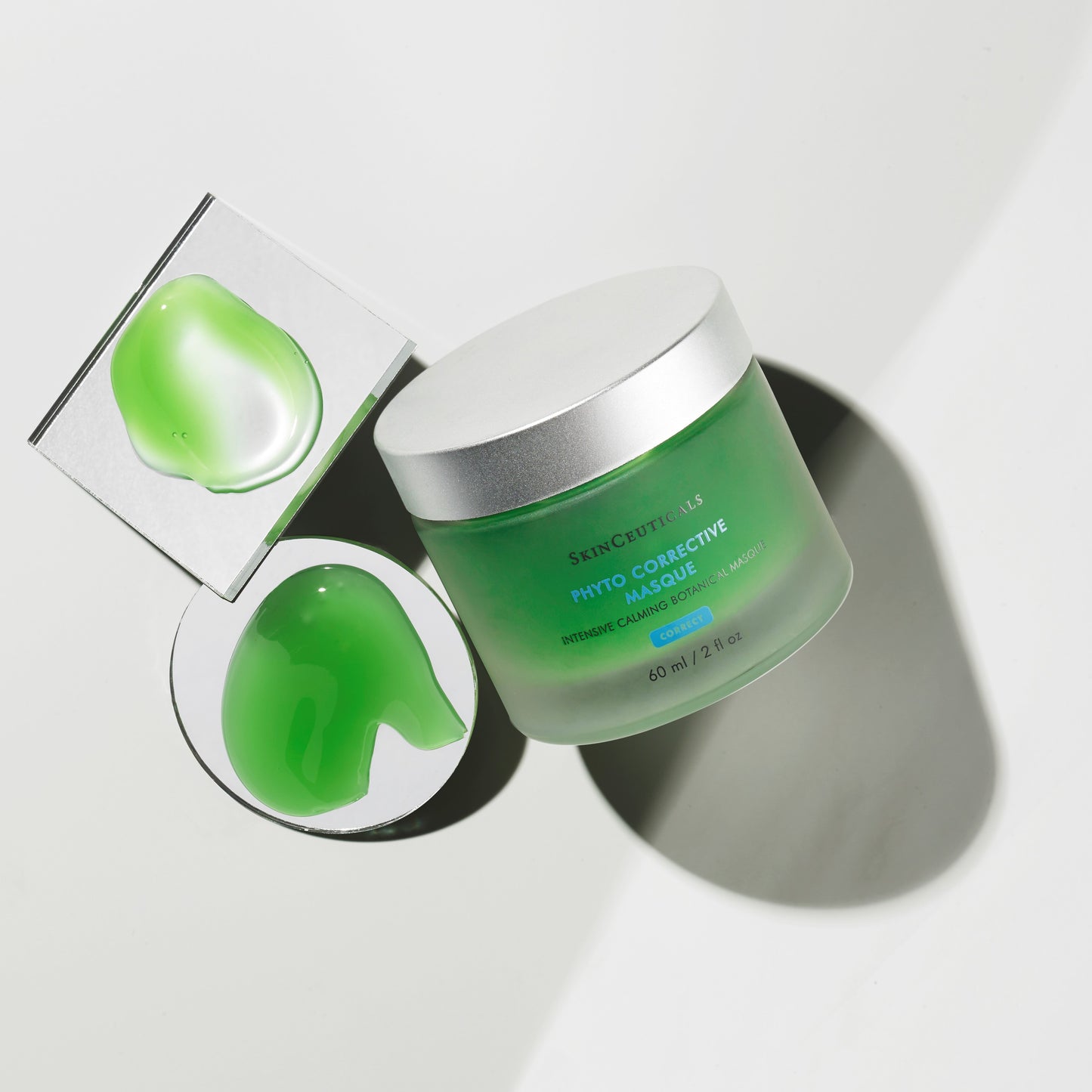 A jar of SkinCeuticals Phyto Corrective Masque with a silver lid is placed next to two green gel samples on reflective surfaces, casting soft shadows on a white background.