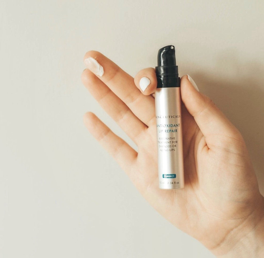 A hand holds a silver bottle labeled "SkinCeuticals ANTIOXIDANT LIP REPAIR" against a plain backdrop, with some product visible on the index finger.